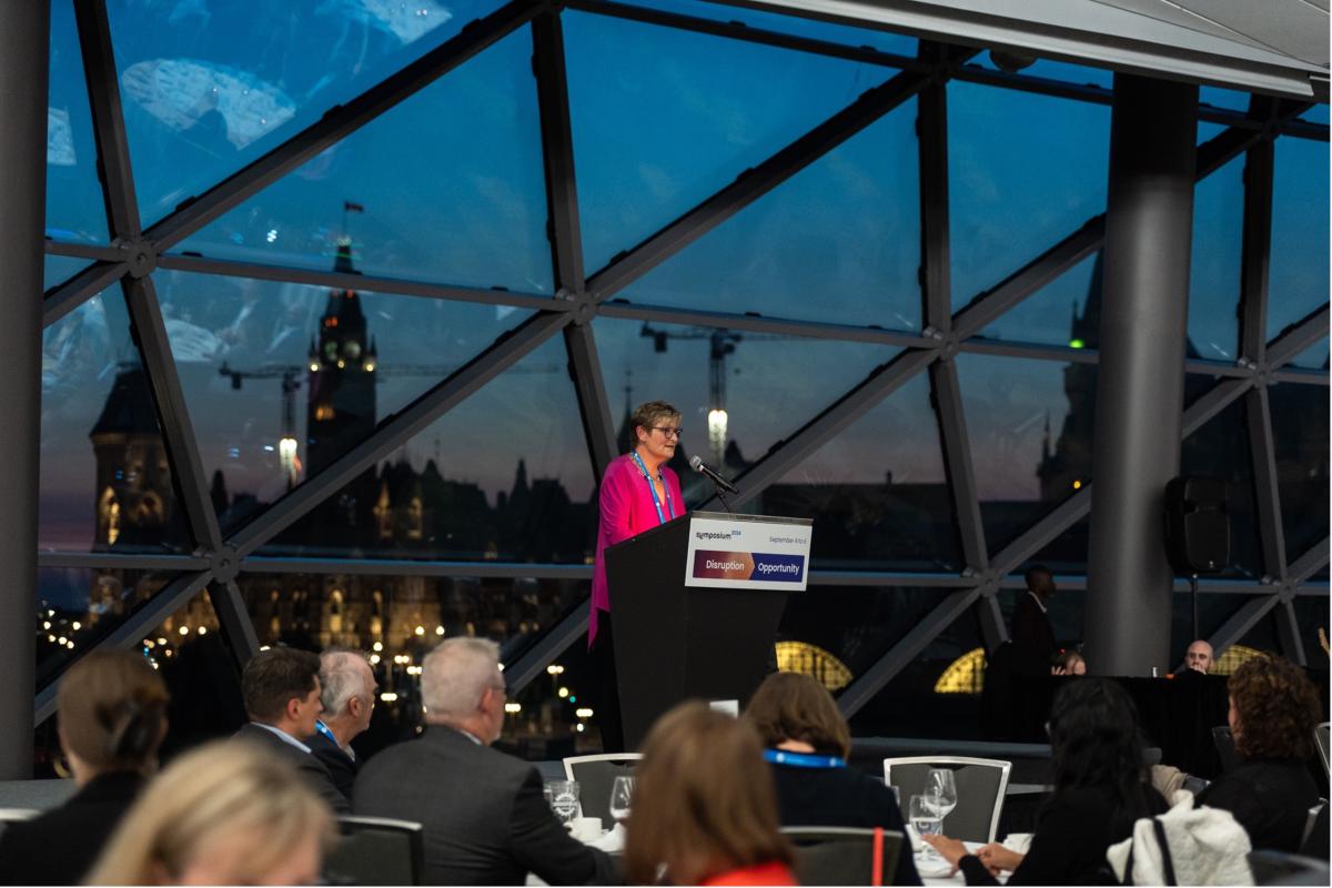 Suzanne McGurn s’adressant aux participants du souper du symposium avec les édifices du Parlement d’Ottawa en arrière-plan au coucher du soleil